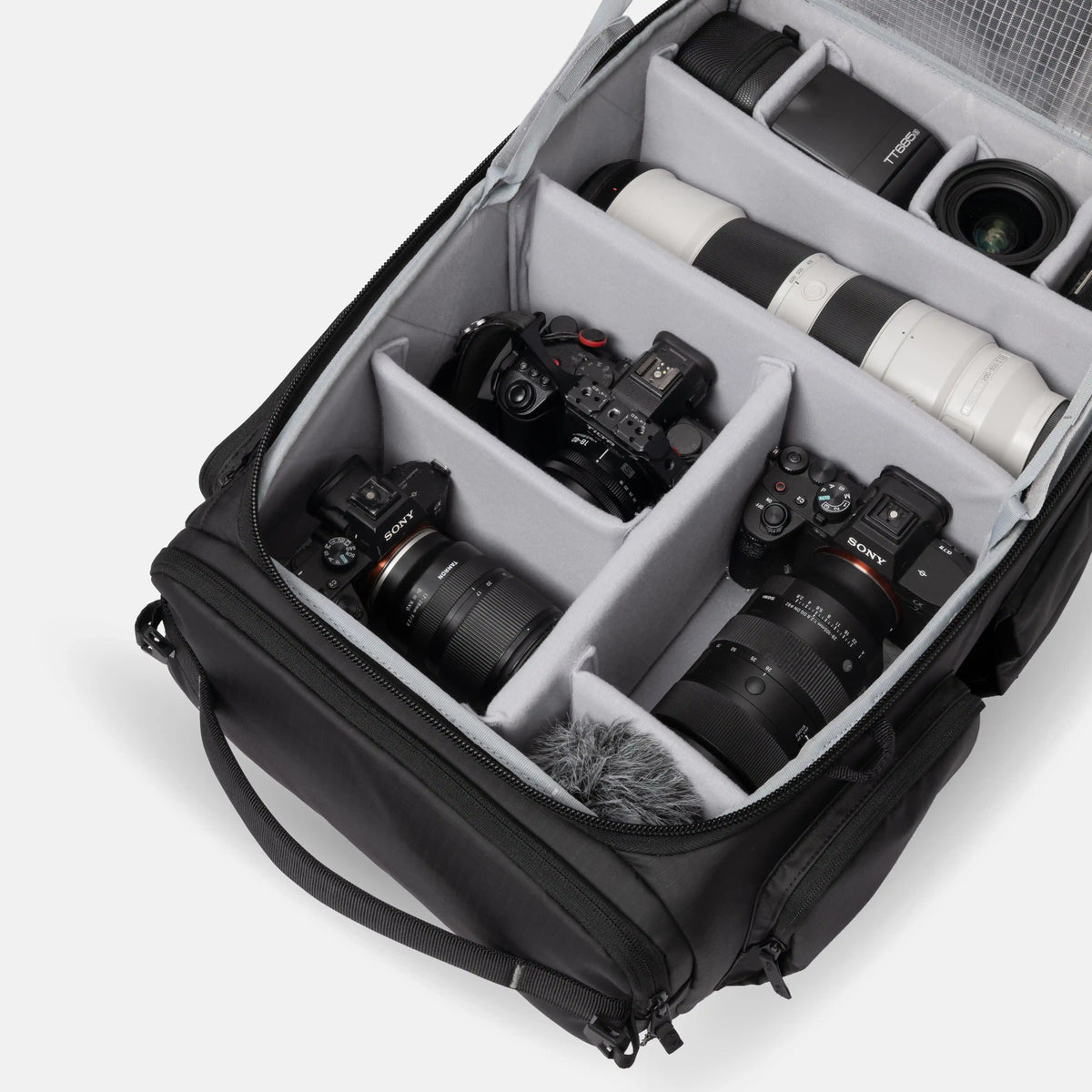 A black camera gear organizer bag shown open, filled with cameras, lenses, and a furry microphone windscreen on a clean white studio background.