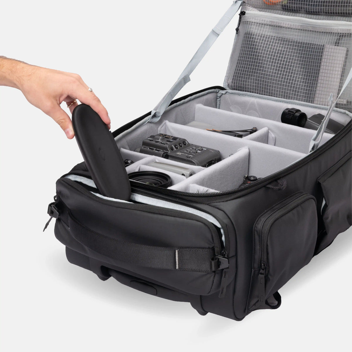 A black camera gear organizer bag with a gray interior organizer system, shown open with a hand placing a black case inside. The bag features multiple compartments, a side pocket, and a mesh lid panel, photographed on a clean light gray background.