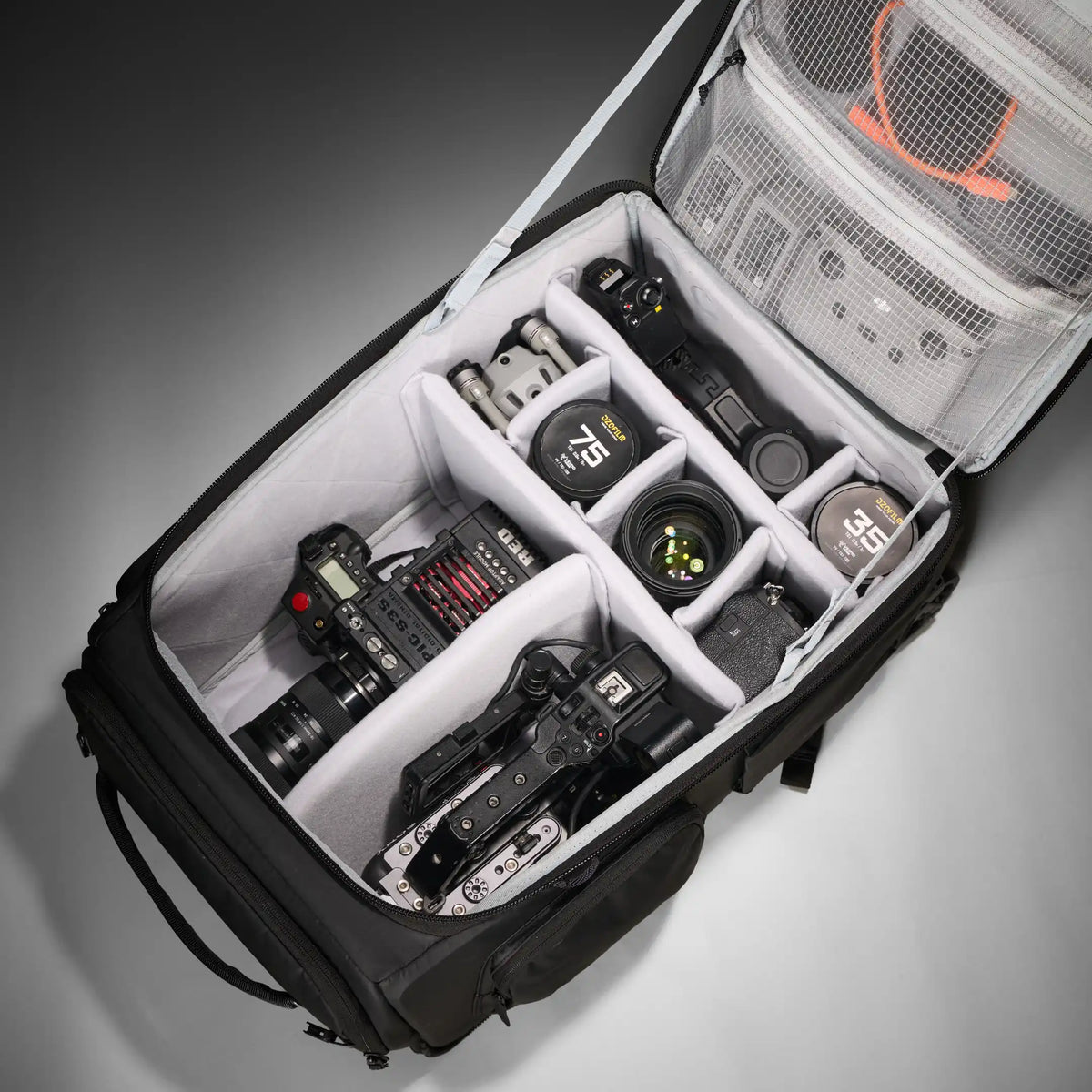 A black camera gear bag shown open from an overhead angle, filled with cameras, lenses, and accessories in white foam compartments and mesh pockets, on a gray studio background.