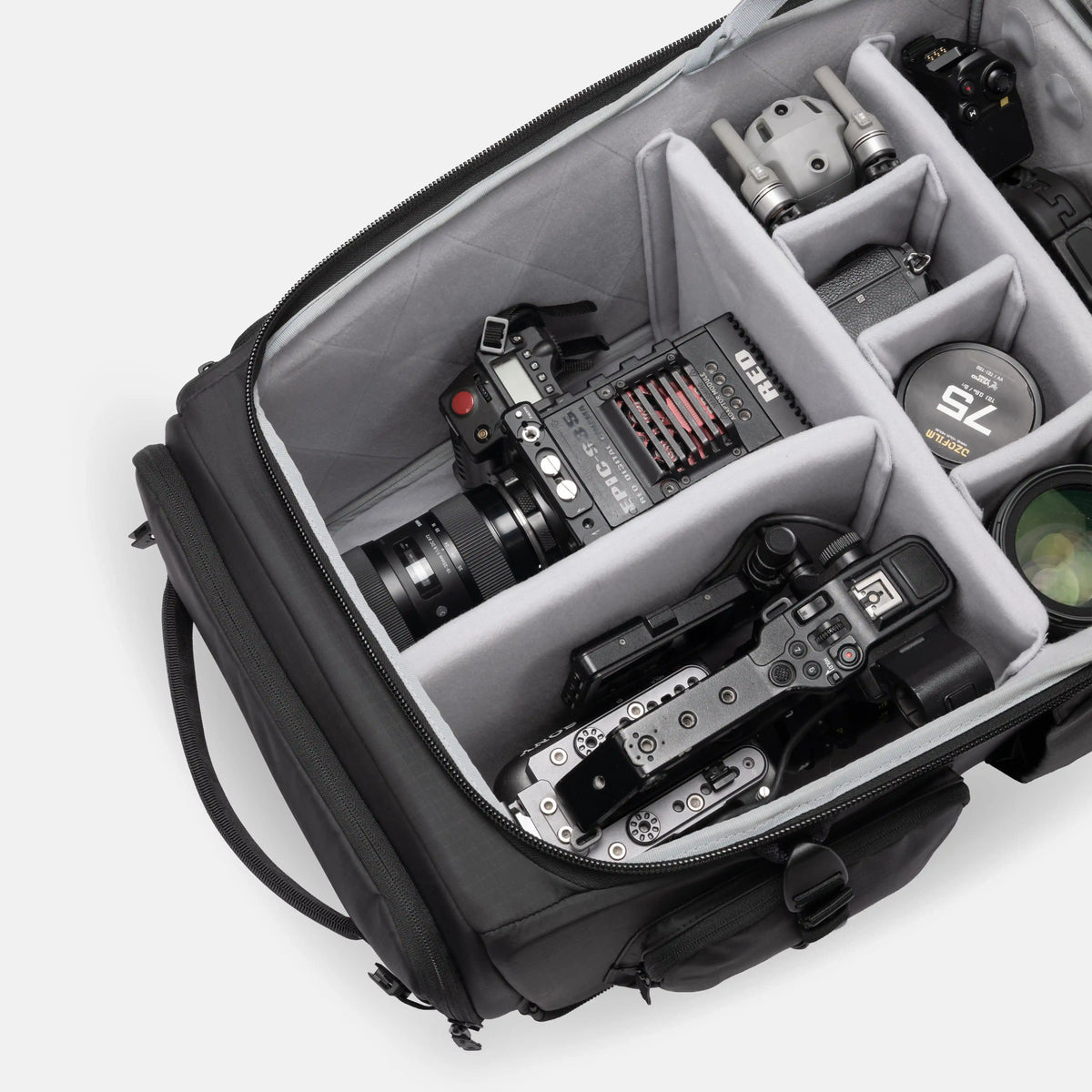 An open black camera backpack with gray foam partition compartments, shown from above and filled with camera gear including a mirrorless camera, lens, accessories, and a drone controller, on a light gray studio background.