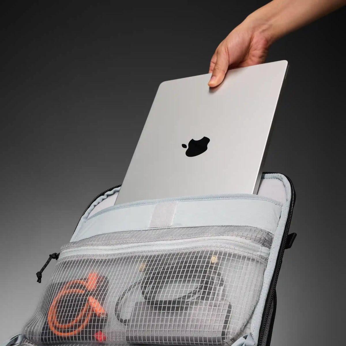 A hand is inserting a silver Apple MacBook into the top compartment of a light gray backpack. The backpack is open, showing a mesh front pocket with an orange cable and other gear inside, set against a dark gray gradient background.