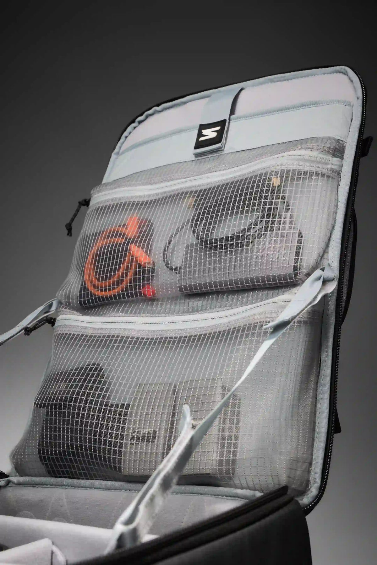 A close-up of an open gray camera gear organizer bag with mesh pockets, showing a camera cable with an orange logo and other gear inside, set against a dark gray gradient background.
