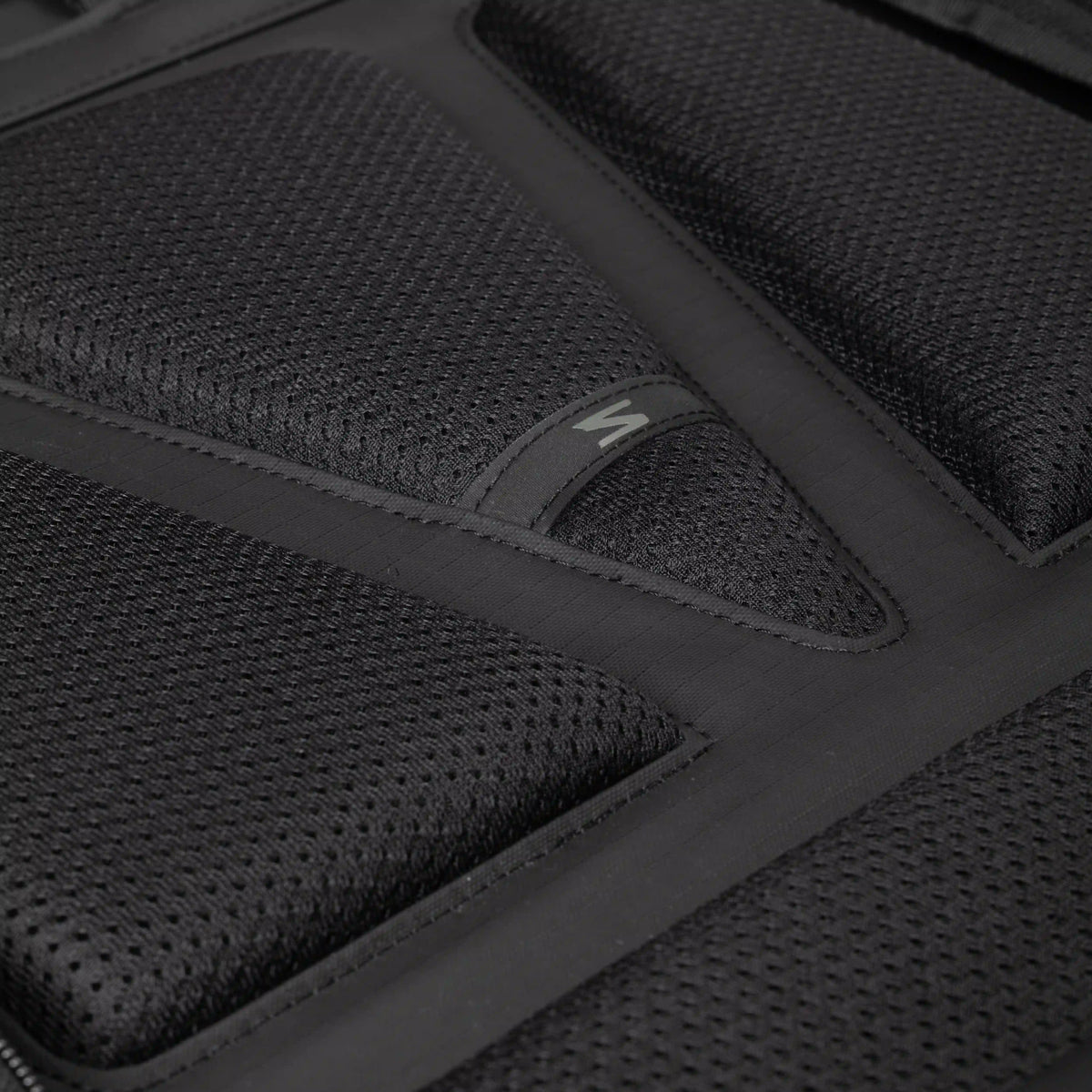 Close-up of a black textured mesh padded surface with stitched panel sections and a small white stylized logo on a black tab.