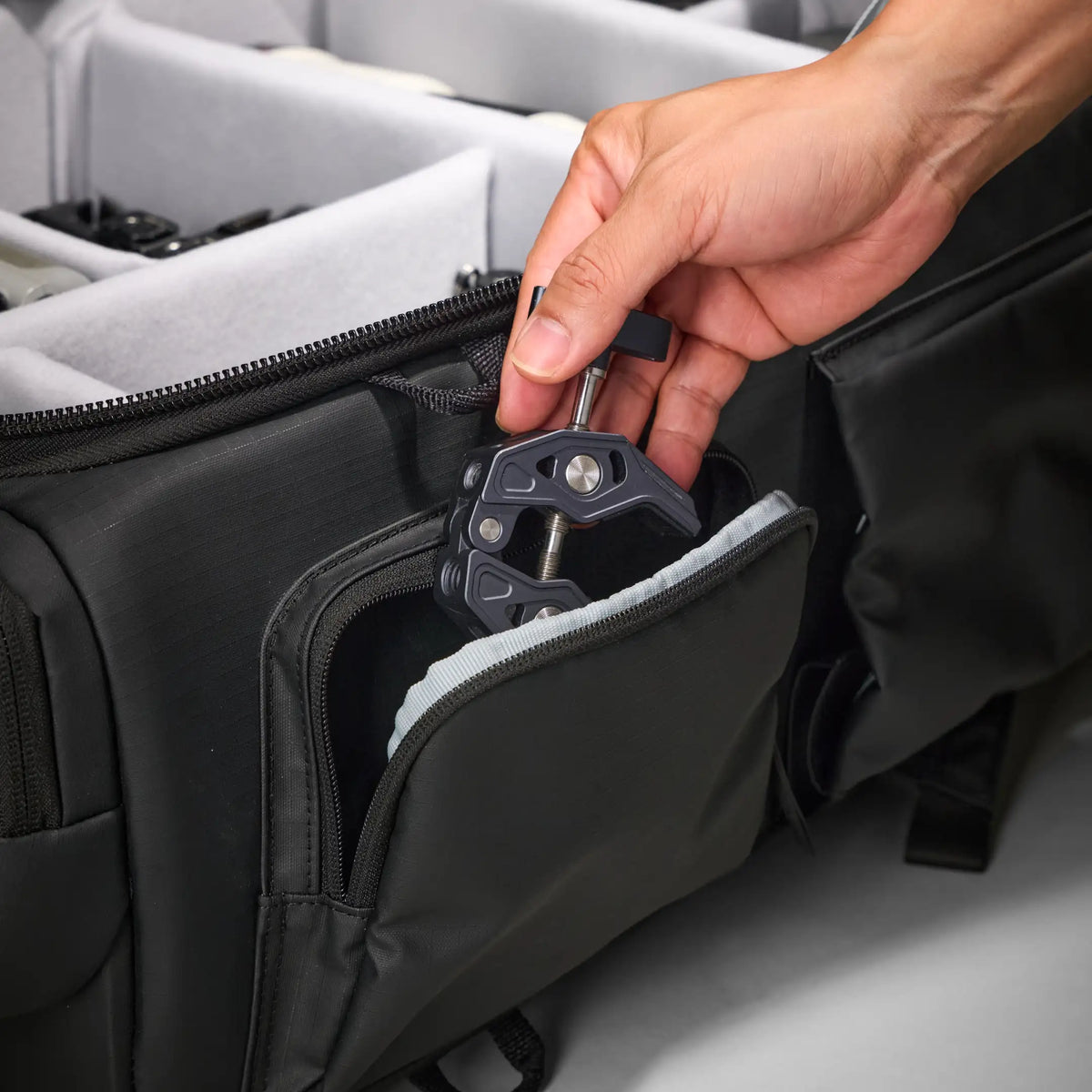 A close-up of a hand inserting a black camera accessory into a zippered pocket of a black camera bag with light gray interior padding.