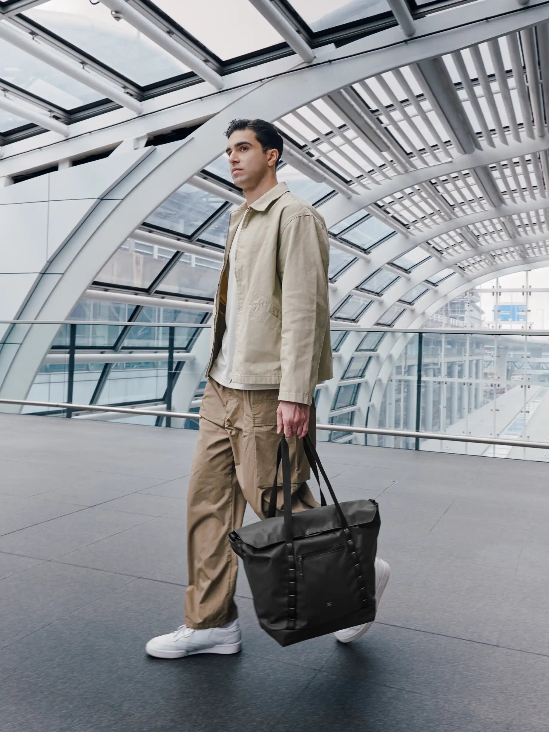 Man in beige jacket and pants holding a black bag in a modern architectural setting