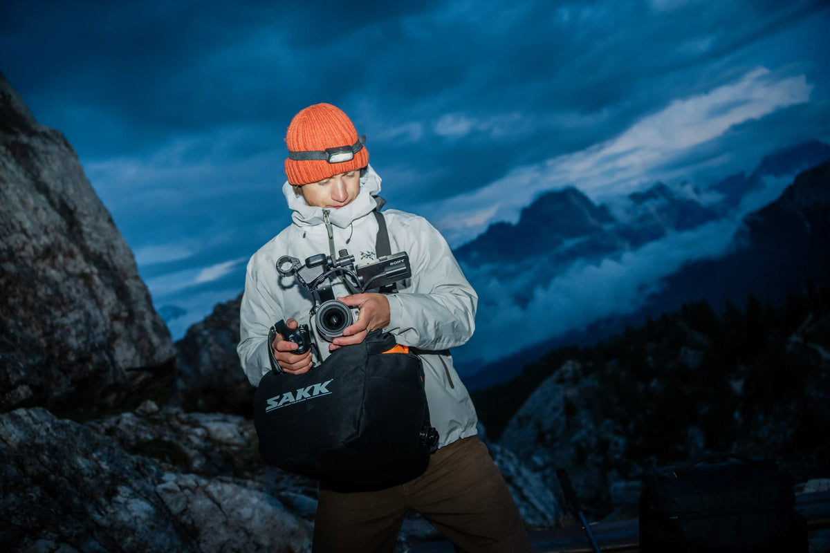 A person in a snowy mountain setting at dusk, wearing an orange beanie and a light gray hooded jacket while holding a camera support bag.