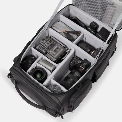 An open black camera gear bag with gray foam compartments, shown from above on a light gray background. The bag is filled with cameras, lenses, a mixer, and other photography accessories.