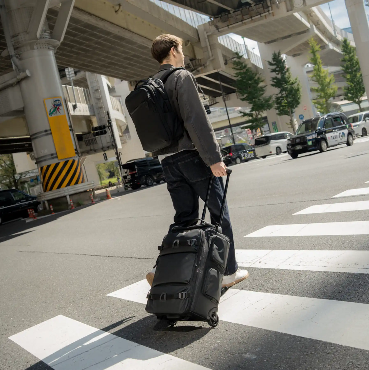 A man wearing a dark gray hoodie and black pants walks across a city crosswalk while pulling a black rolling suitcase with a telescoping handle. He is also wearing a black backpack.