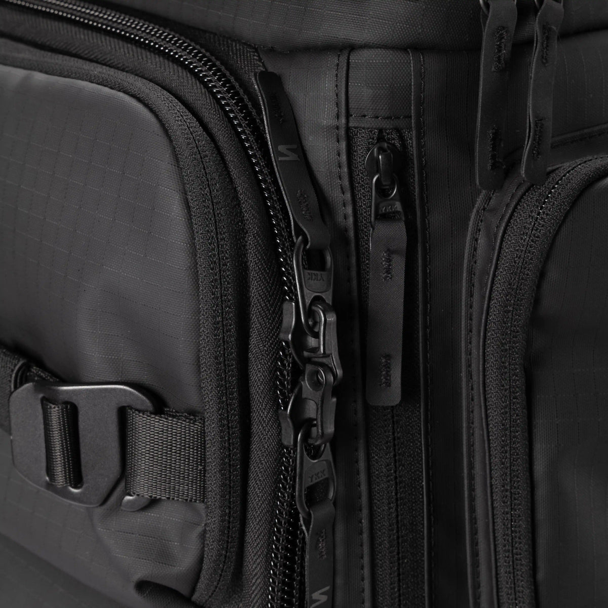 Close-up detail of a black technical backpack showing multiple zippered compartments, a buckle strap, and textured fabric.