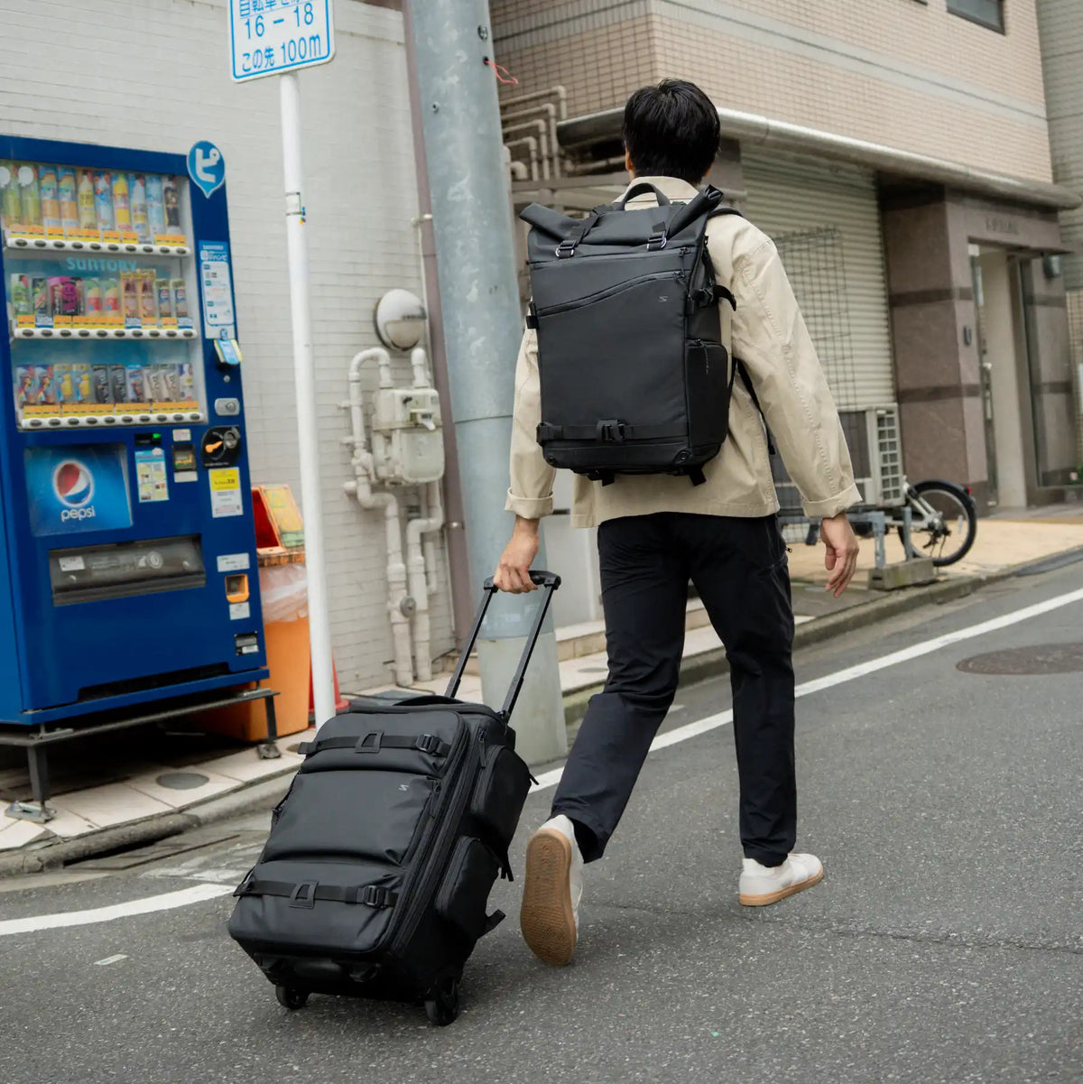 A man seen from behind walking on a city street, wearing a black backpack and pulling a black rolling suitcase. The scene includes a blue vending machine, building walls, a utility pole, and a bicycle in the background.