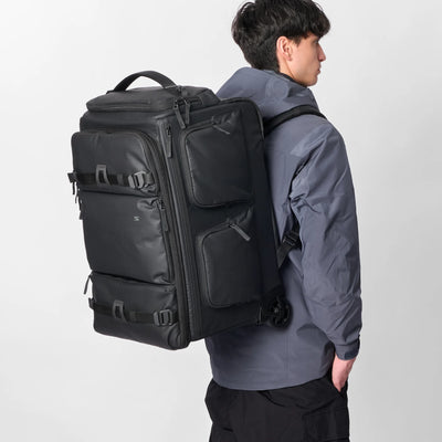 A man wearing a dark gray jacket and black pants is shown from the back wearing a large black rolling travel backpack with multiple zippered compartments and side straps, set against a plain light gray studio background.