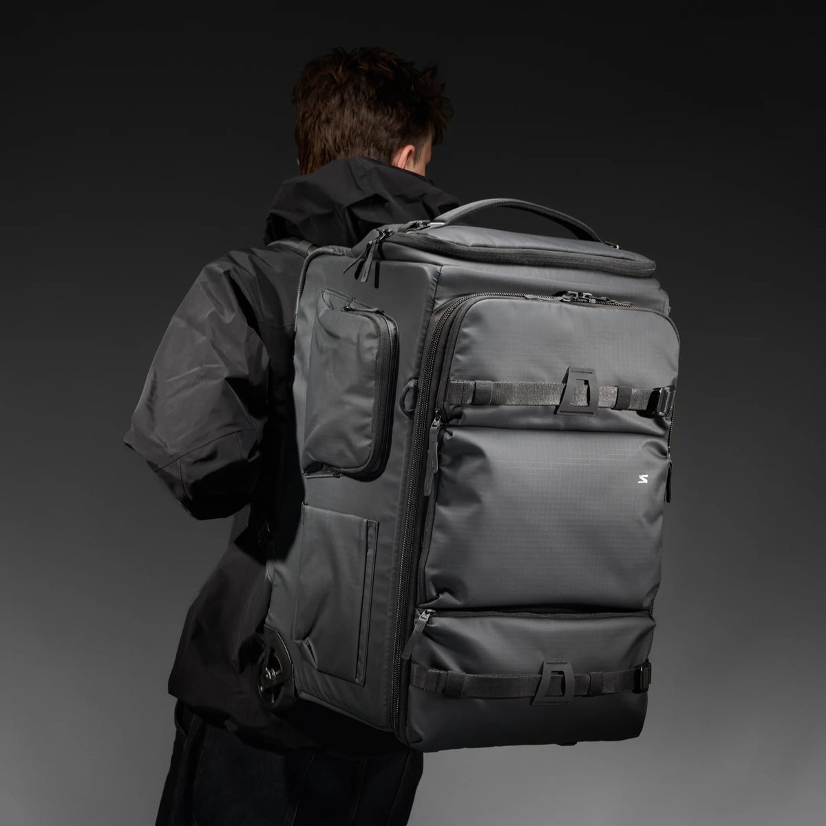 A man viewed from behind wearing a large dark gray rolling duffel backpack with multiple zip compartments and side pockets, set against a dark gray studio background.