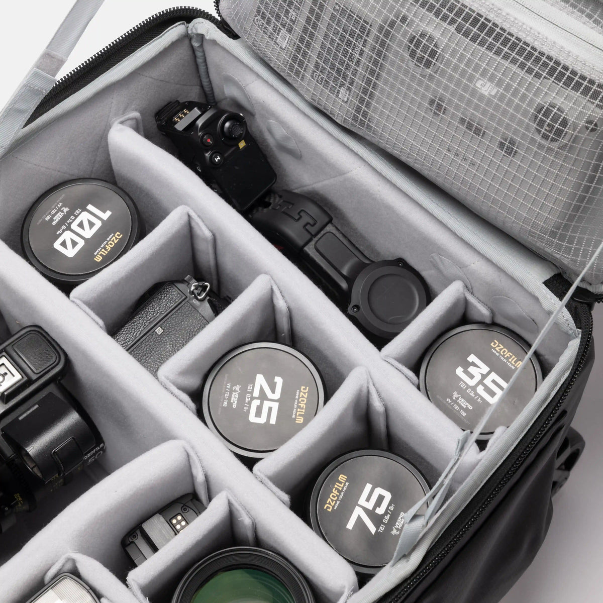 An open black camera gear bag with gray foam compartments, shown holding camera lens caps and camera accessories. The bag's interior organizer is clearly visible, along with several lens caps featuring gold and white markings.