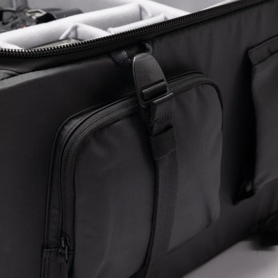 Close-up of a black soft-sided travel or equipment bag with a front zippered pocket and an adjustable hanging strap, shown against a light neutral background.