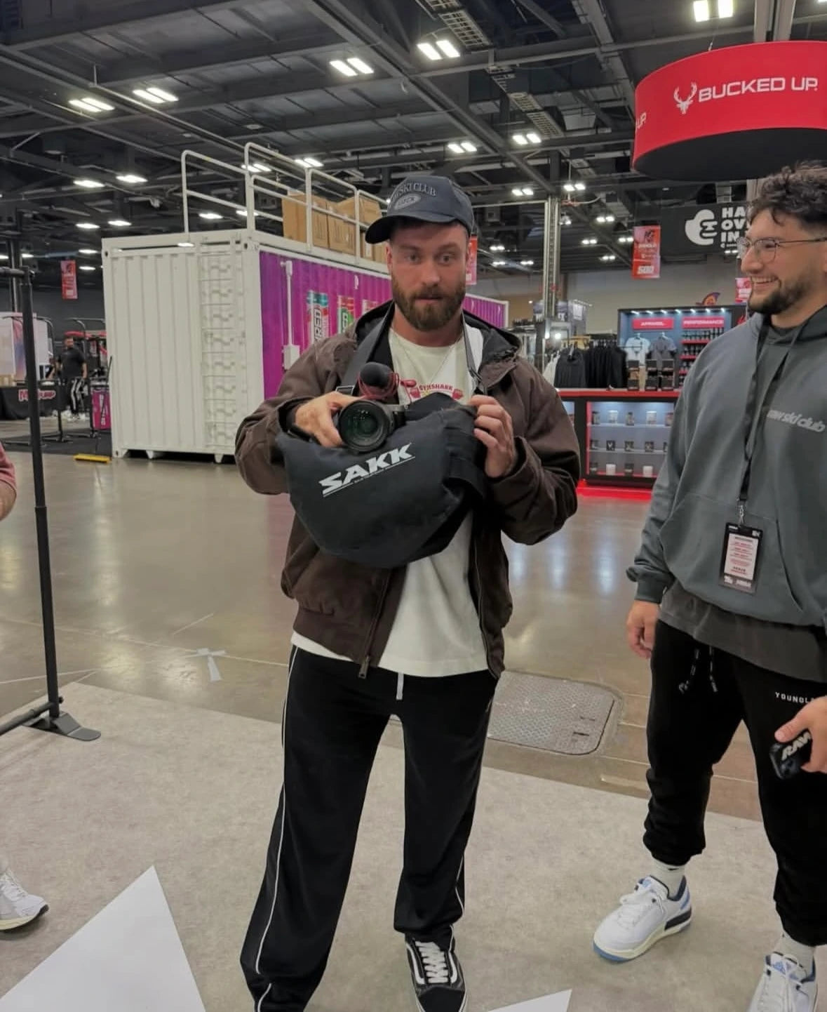 Person holding a Sakk Gear camera bag at an indoor event while another person watches.