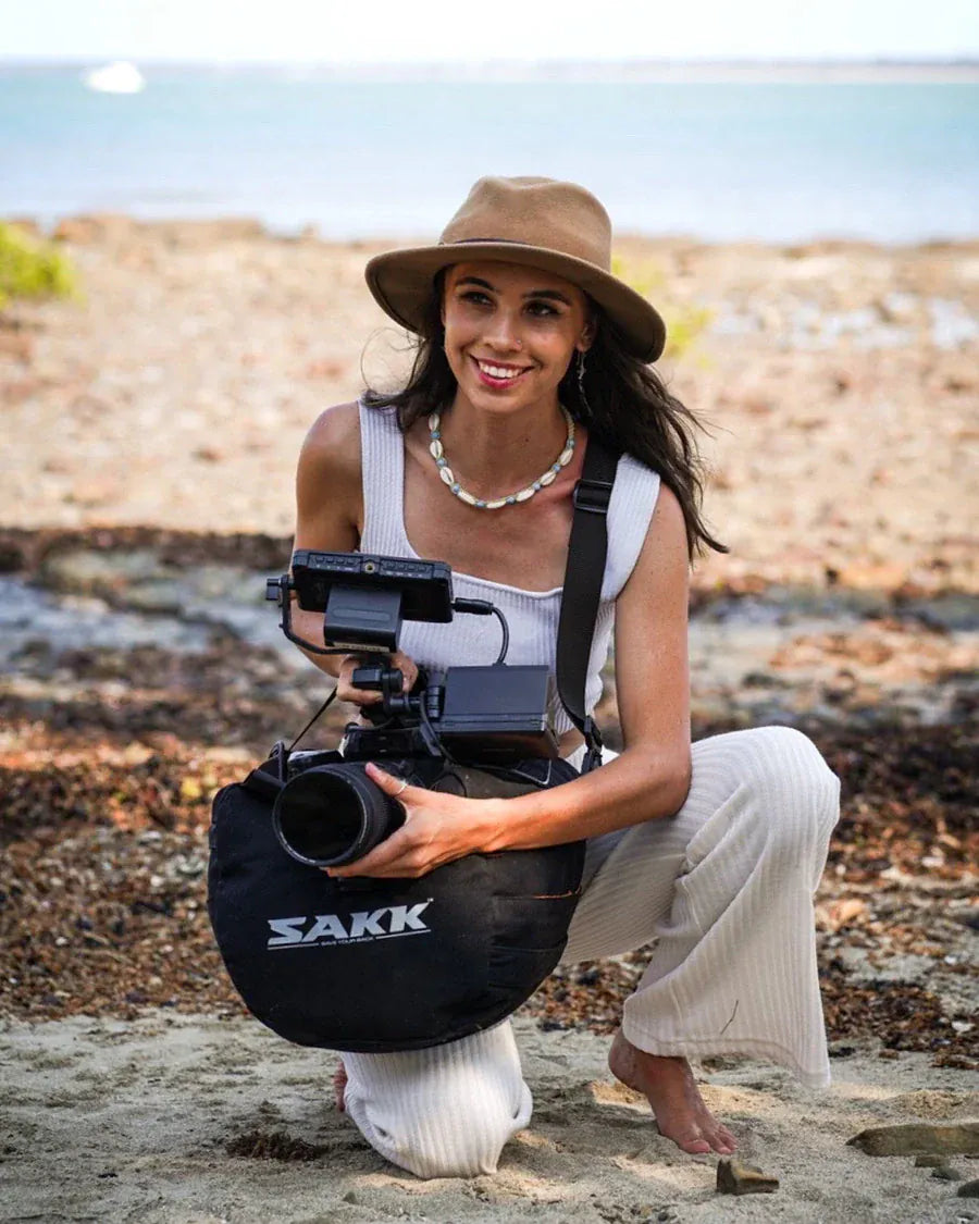 Woman wearing a brown hat and white outfit kneels barefoot on a beach, using a Sakk Gear bag to support her professional camera rig while smiling at the camera