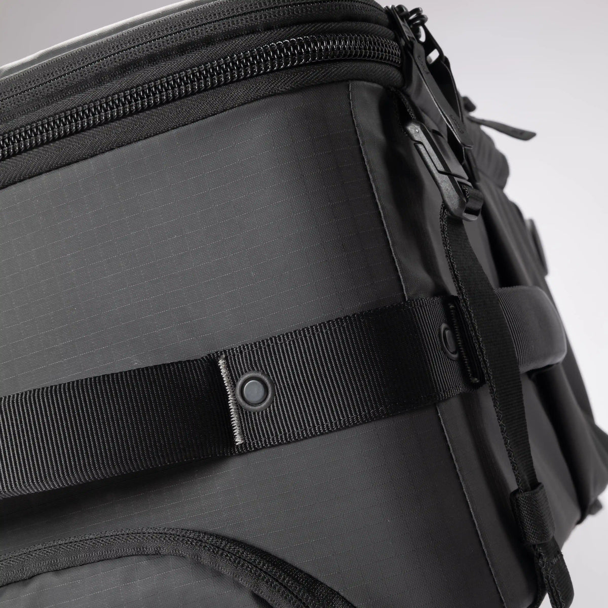Close-up of a black technical backpack showing the textured ripstop fabric, a black webbing strap with a snap button closure, and a zipper compartment.