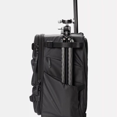 A black soft-sided travel or camera bag with a tripod mounted vertically inside a side pocket, shown against a plain light gray studio background.