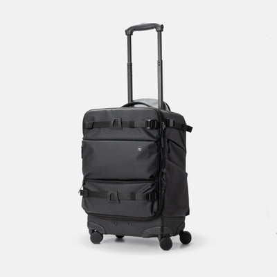 A black rolling suitcase with an extended telescoping handle, spinner wheels, and multiple compression straps on the front, shown against a light gray studio background.