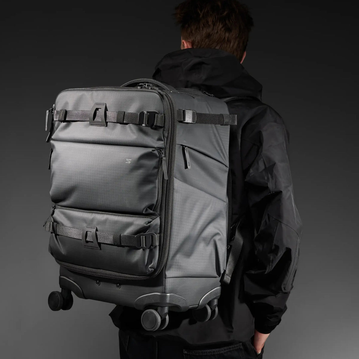 A person viewed from behind wearing a large dark gray rolling backpack with multiple compression straps and spinner wheels, over a black hooded jacket, against a dark studio background.
