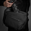 Person holding a camera with a lens attached to a black shoulder bag on a dark background