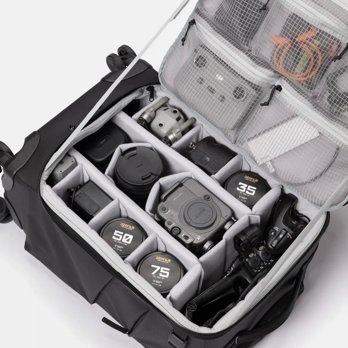 An open black camera gear bag with gray interior organizer compartments, shown on a white studio background. Visible items include lens caps labeled Sigma 35mm, 50mm, and 75mm, a camera body, a lens, and mesh pockets with accessories.