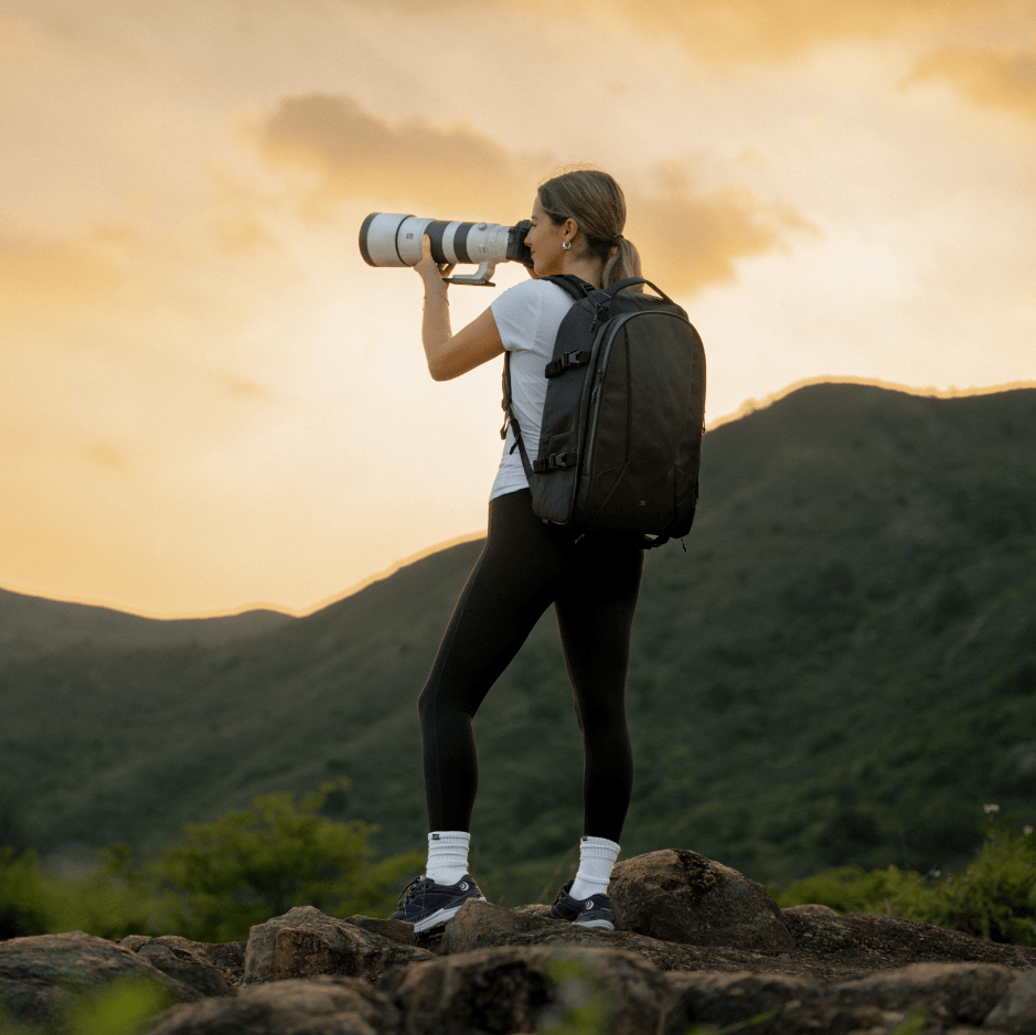 Person with camera and backpack taking a photo of a mountain landscape at sunset.
