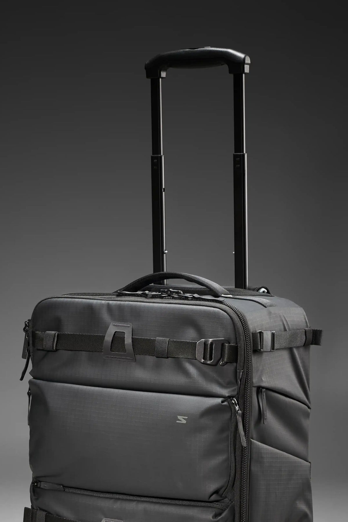 A dark gray rolling duffel bag with an extended black telescoping handle, shown against a dark gray gradient studio background.