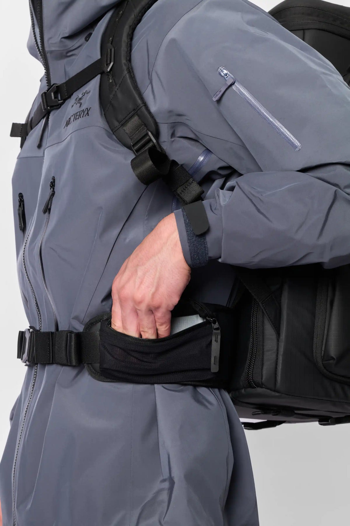 Close-up of a person wearing a gray Arc'teryx jacket and black backpack, with the hand reaching into a side zippered pocket of the jacket.