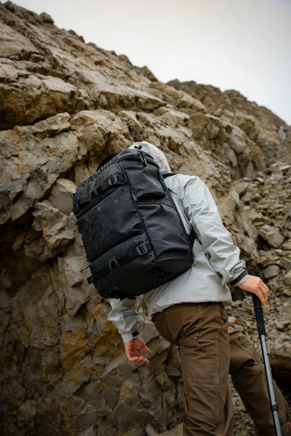 A hiker wearing a large black waterproof backpack, a light gray jacket, and brown pants navigates a rocky mountain path, holding a trekking pole.