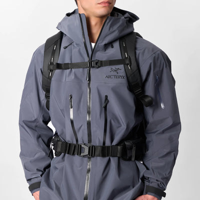 A man wearing a gray hooded technical jacket with black harness straps and a black backpack, shown from the chin down against a light gray studio background.